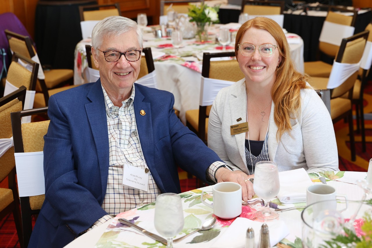 Two people sit at a table with glasses and mugs at an event.