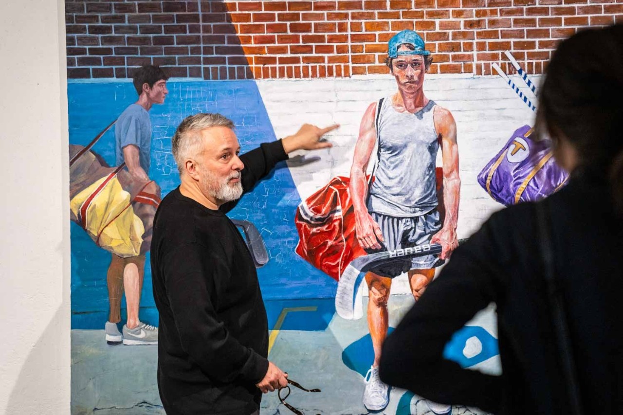 McMinn pointing to one of his paintings of a young street hockey player as someone looks on.