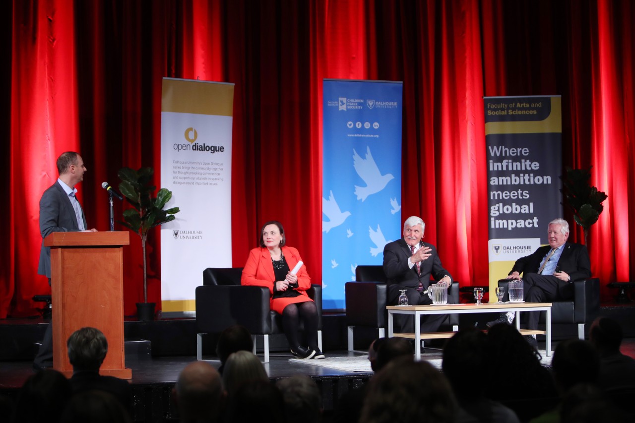 Three people sit on stage with banners red curtains in the background while another person stands at the podium addressing them.