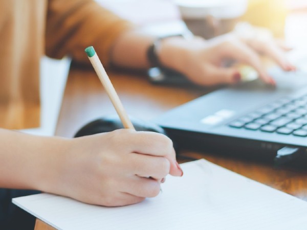 A close-up of a person holding a pencil to paper in one hand with the other hand on a laptop. 