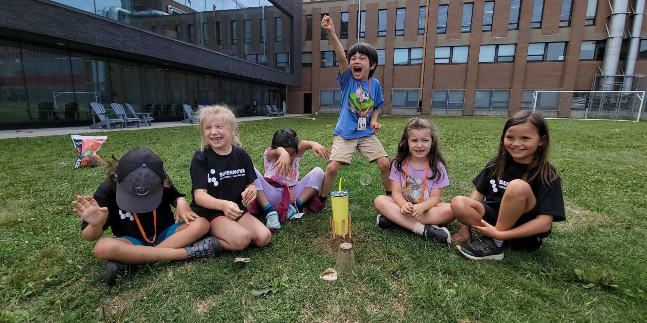 Children sitting on grass with a small wooden structure in the center. One child is standing, excitedly raising a hand. A building is in the background.