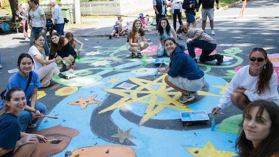 A group of young people sit on the street painting a brightly coloured mural.
