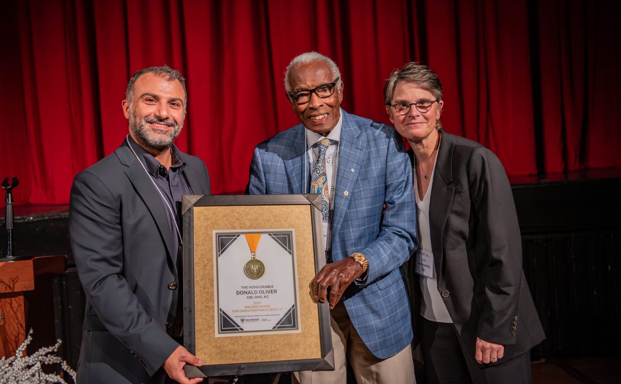 Senator Oliver holds a framed certificate and stands between two people against a stage backdrop with a red curtain.