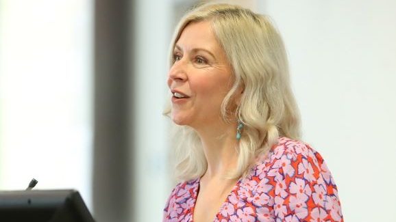 A close-up of Dr. Anna MacLeod, RIM Unit Head, speaking at a podium, looking towards an audience.