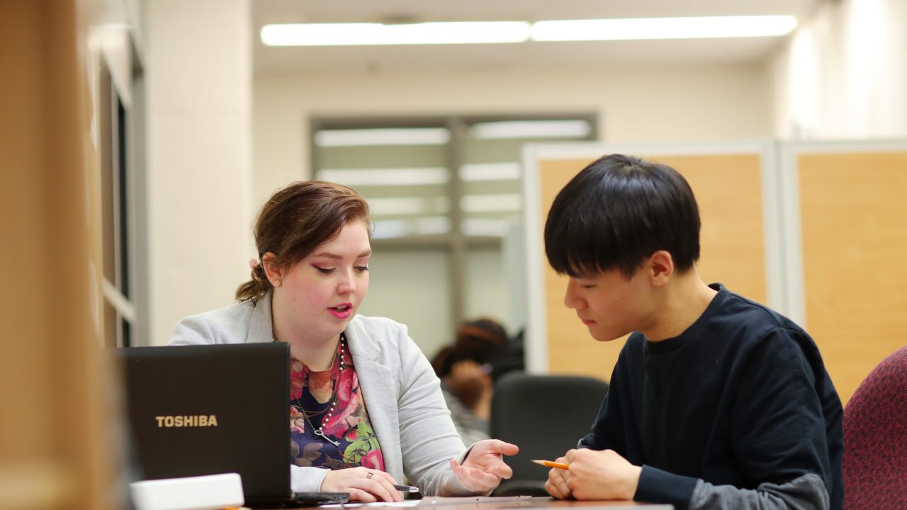 A female-presenting person assists a male-presenting person while seated at a table.