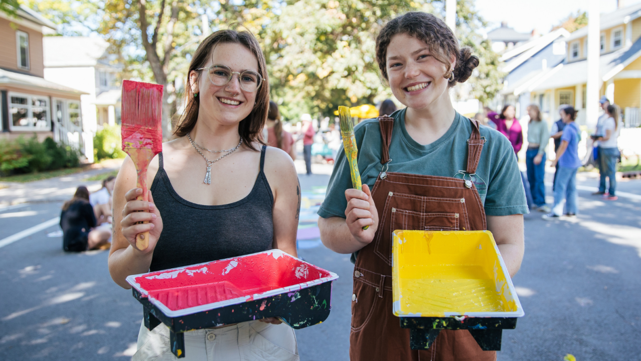two female students holding paintbrushes and paint trays, laughing